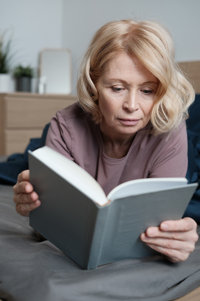 An elderly woman with short blond hair in light house clothes reading in bed