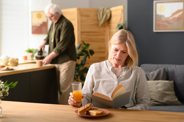 An aged woman with short blonde hair reading a book and having breakfast at home