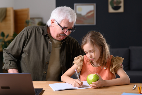 An elderly man in glasses helping his granddaughter with a green apple in her hand to do homework