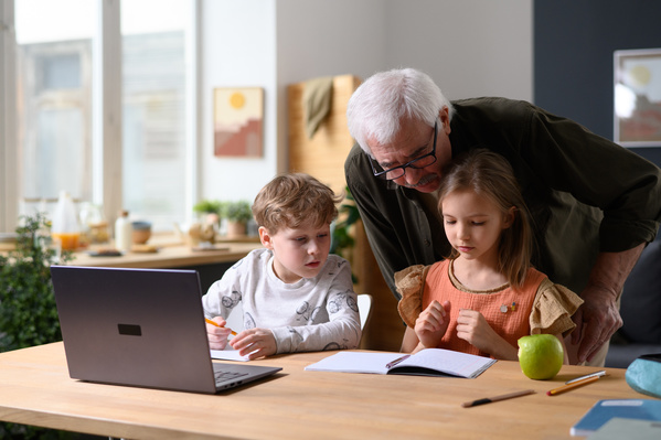 An elderly man in glasses helping his grandchildren do their homework at a wooden table with a laptop