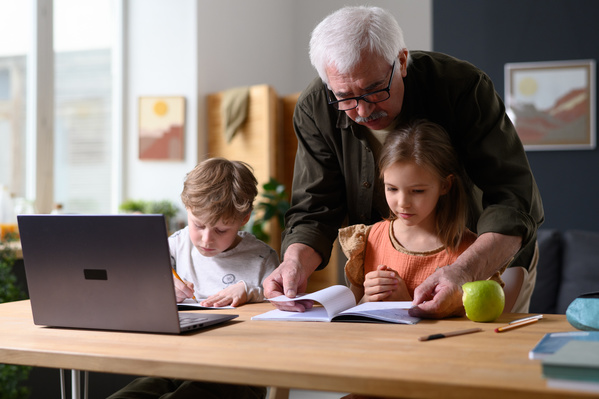 A grandpa in glasses helping his grandchildren to do homework at a wooden table with a laptop