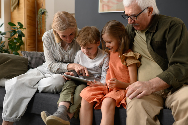 Elderly grandparents with blonde hair and their grandchildren using a tablet on a dark sofa