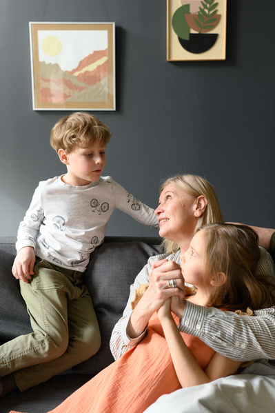 A blond-haired boy chatting with his elderly grandmother and little sister sitting on a dark sofa