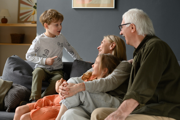 A blond-haired boy chatting with his elderly grandparents and little sister sitting on a dark sofa