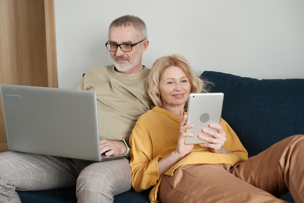 An elderly man and a woman with blonde hair using gadgets on a dark blue sofa at home