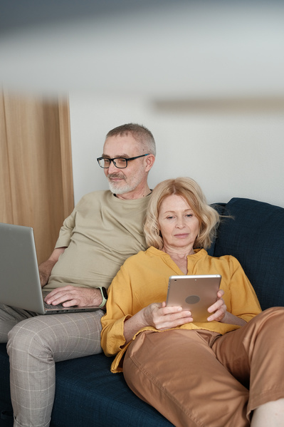An elderly couple with blonde hair using gadgets on a dark blue sofa at home