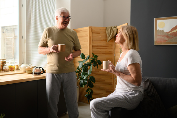 A gray-haired man in pajamas chatting over a cup of tea with his wife in a white T-shirt and light pants
