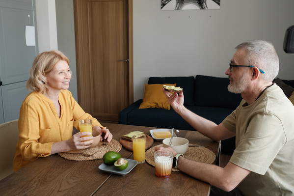 An elderly husband and wife in light outfits chatting during breakfast in a bright kitchen
