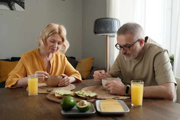 An elderly wife with blonde hair and her husband in a light T-shirt eating porridge in a bright kitchen