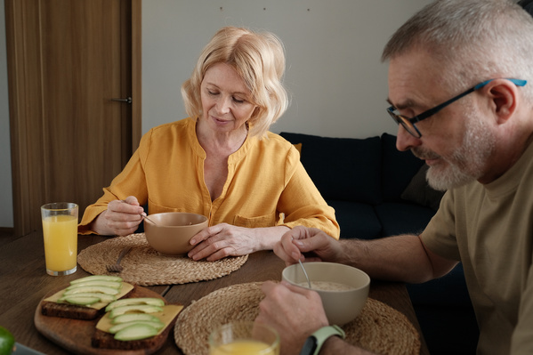 An elderly wife with blonde hair and her husband in a light T-shirt having breakfast in a bright kitchen