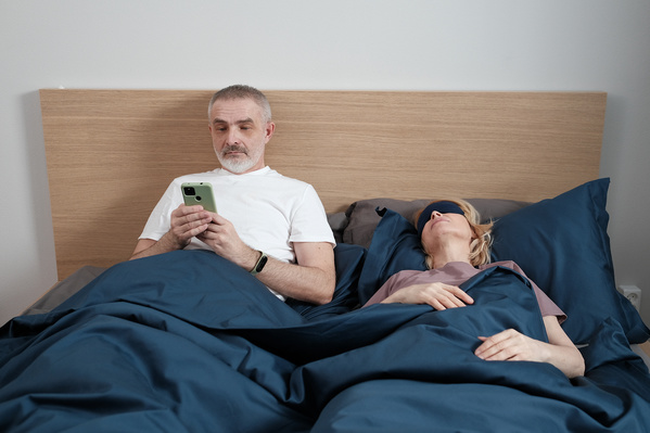 A man in a white T-shirt using a phone in bed with his sleeping wife