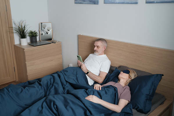 A man in a white T-shirt surfing Internet on his phone and his sleeping wife with a mask over her eyes in bed