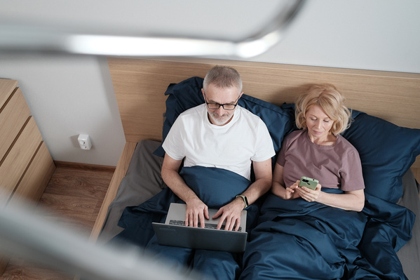 An elderly man and woman in light pajamas using gadgets in bed before going to bed
