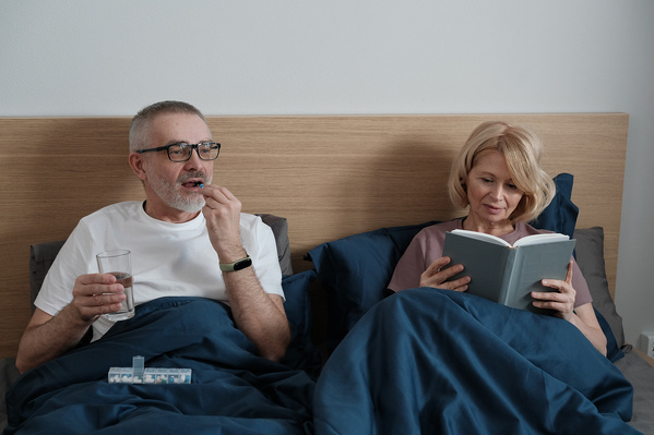 A blonde elderly woman reading in bed and her husband taking a pill next to her