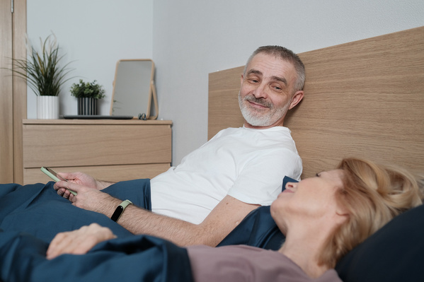 A man in a white T-shirt with a phone smiling at his blonde wife in bed