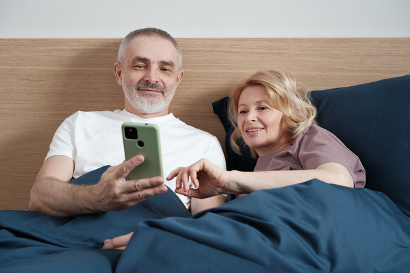 An elderly husband and wife in light pajamas using the phone while lying in bed