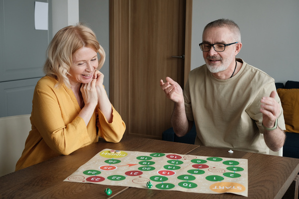 An elderly smiling wife with blonde hair and her husband in a light T-shirt playing a bright board game