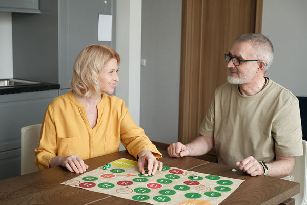 An elderly woman and her husband with glasses playing a bright board game at a wooden table in a bright room