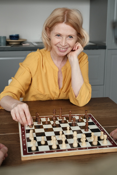 An elderly smiling woman playing chess at a wooden table in a bright room