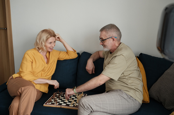 An elderly man making a move while playing chess with his blonde wife at a wooden table in a bright room