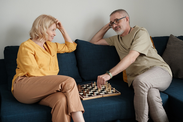 An elderly couple in light outfits playing chess on a dark blue sofa