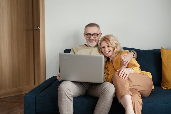 An elderly husband and wife in light outfits watching a movie on a silver laptop while sitting on a blue sofa