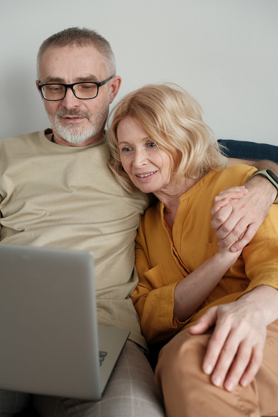 An elderly couple in light outfits watching a movie on a silver laptop while sitting on a blue sofa