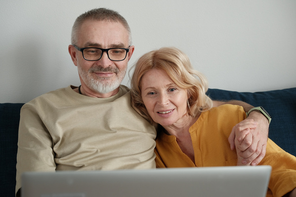 An elderly man and woman in light outfits watching a movie on a silver laptop while sitting on a blue sofa