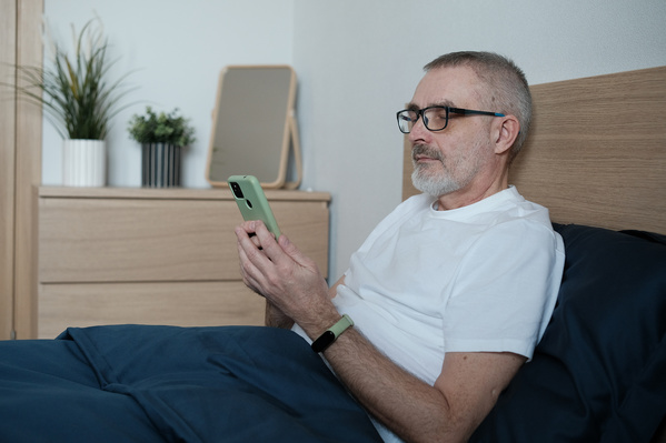 An aged man with gray hair in a white T-shirt using a phone in bed before going to sleep