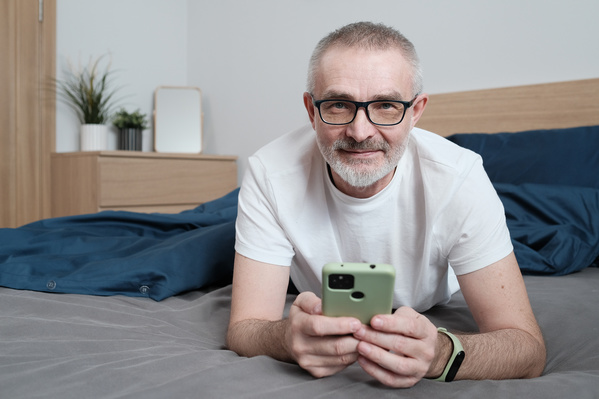 An elderly man with glasses lying in bed with a phone in the morning