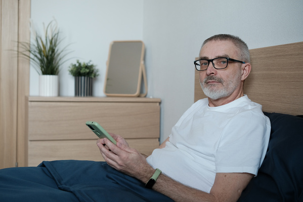 An aged man with glasses sitting in bed with a phone in the morning