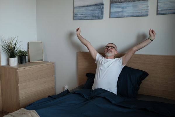 An aged man in glasses and a white T-shirt stretching in bed in the morning