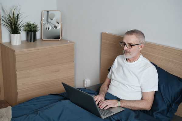 An aged man with a beard dressed in home clothes using a laptop in bed
