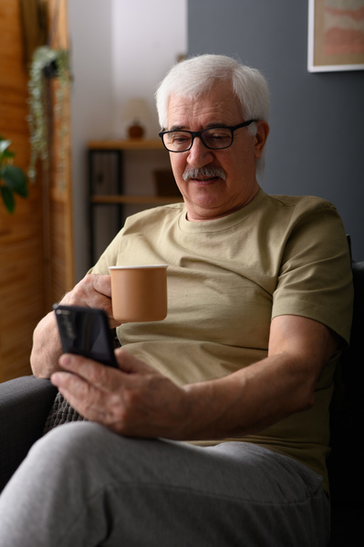 An elderly man in glasses watching a video on his phone over a cup of tea while sitting on a gray sofa