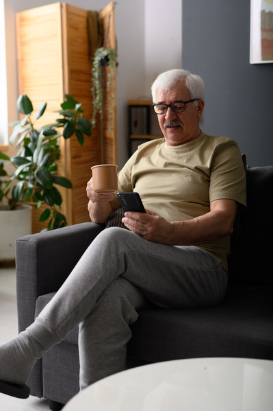 An elderly man in glasses surfing the Internet on his phone over a cup of tea while sitting on a gray sofa