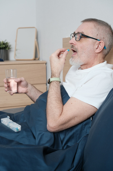 An aged man in glasses and light clothes taking a pill in bed