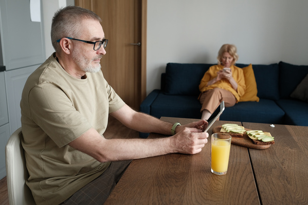An elderly man in glasses and pajamas using a tablet at breakfast