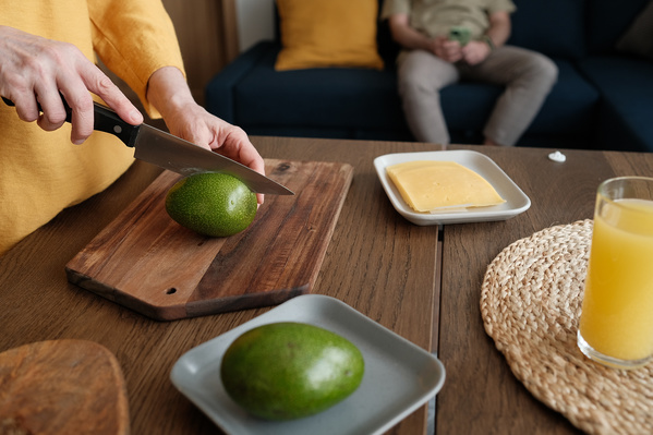 A woman in a yellow blouse cutting an avocado in half on a wooden chopping board for breakfast