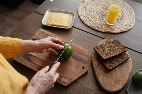 A woman in a yellow blouse slicing an avocado in half on a wooden chopping board for breakfast