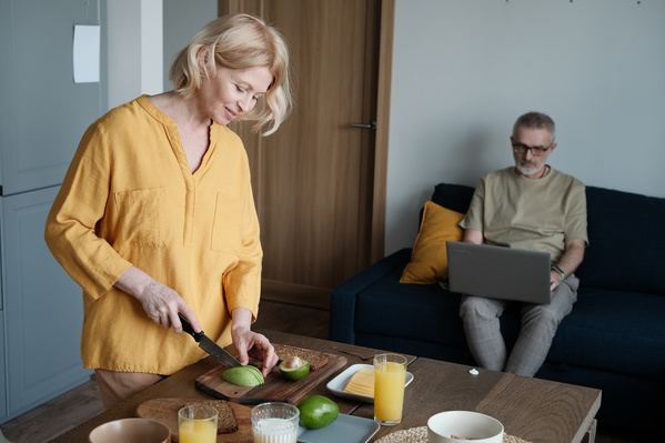 A senior woman with short blonde hair in a yellow blouse cutting an avocado on a wooden chopping board for breakfast