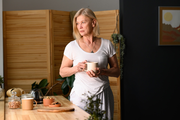 A senior woman with short blond hair in light house clothes drinking tea in the kitchen