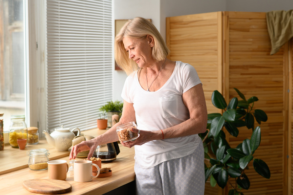 A blonde elderly lady in light pajamas making tea in the kitchen