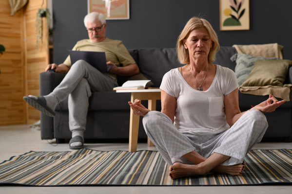 An elderly woman with short blond hair in light home clothes practicing yoga at home
