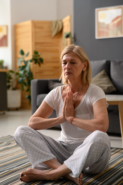 An aged woman with short blond hair in light home clothes practicing yoga at home