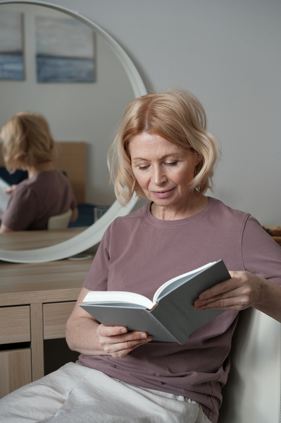 An aged woman with short blonde hair dressed in pajamas reading a book in the bedroom