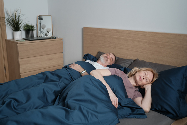 An elderly man in light pajamas sleeping with his wife in a dark blue bed