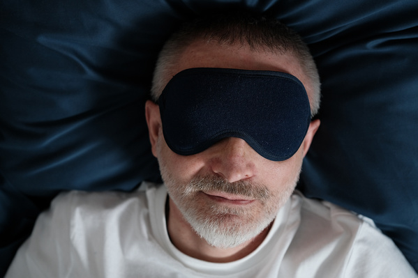 An elderly man in a white T-shirt sleeping in a dark blue eye mask