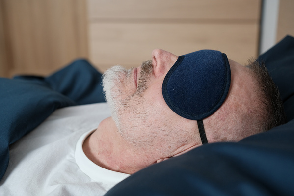An elderly man with a beard in a white T-shirt resting in a dark blue sleep mask