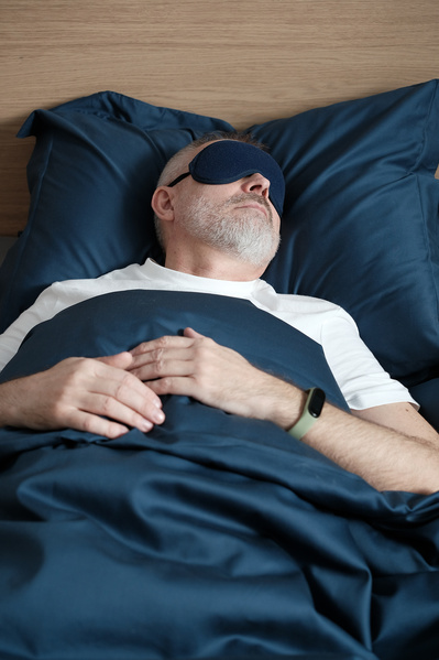 An elderly man with a beard in a white T-shirt relaxing in a dark blue sleep mask
