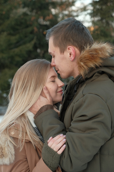 A man in warm clothes kissing a blonde woman on the forehead in a winter forest
